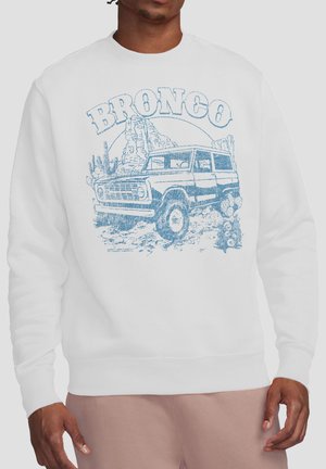 Man wearing white sweatshirt with blue vintage Bronco SUV graphic and desert landscape with cacti and rock formations.