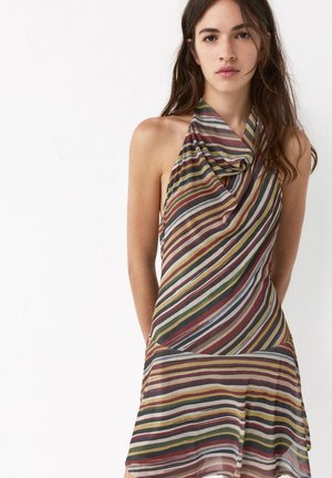 Young woman wearing a sleeveless, multicolored striped halter dress standing against a plain white background.