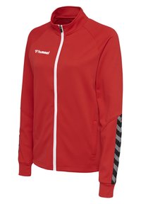 Rode sportjas met hoge kraag, volledige witte rits, textuurstof en zwarte chevronstrepen op de mouwen. Hummel-logo in het wit.