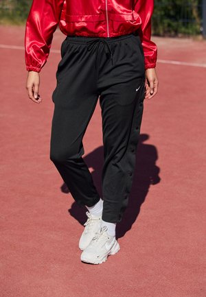 Osoba w czarnych spodniach dresowych Nike, białych tenisówkach i czerwonej satynowej kurtce stoi na czerwonym zewnętrznym boisku sportowym.