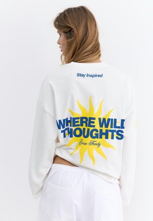 Donna con capelli castano chiaro indossa una felpa bianca con un sole giallo e testo blu "WHERE WILD THOUGHTS" e testo più piccolo "Stay Inspired" e "Grow Freely".