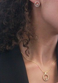 Collana d'oro con pendente circolare e gemme trasparenti; orecchini a bottone coordinati. Capelli ricci e cima nera visibili.