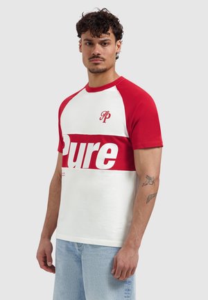 Mann mit lockigem Haar, der ein rot-weißes kurzärmeliges Hemd mit dem Schriftzug „Pure“, hellblaue Jeans und Tätowierungen am rechten Unterarm trägt.