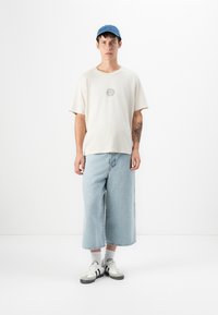 Man in blue cap, beige sun-print t-shirt, light blue wide cropped jeans, white socks, and white sneakers stands against white background.
