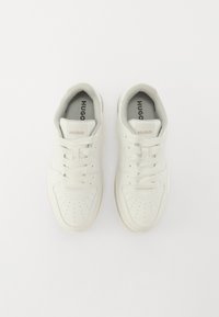 White leather sneakers with rounded toe and textured accents, featuring perforations and six eyelets for laces, with "HUGO" branding on the tongue.