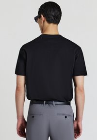 Uomo che indossa una maglietta nera, pantaloni grigi e una cintura nera, di spalle con capelli corti scuri e occhiali da sole neri su uno sfondo chiaro.