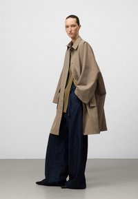 Kobieta z związanymi włosami, ubrana w oversized beżowy płaszcz, oliwkowy top i szerokie ciemne dżinsy, stojąca na jednolitym tle.