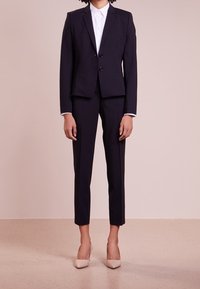 Marineblauw op maat gemaakt pak met een getailleerde blazer en slanke broeken, gecombineerd met een witte blouse met kraag en beige hoge hakken. Zachte stof textuur.