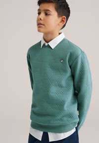 Zelený sveter s hexagonálnym vzorom vyrobený z textúrovanej látky, so zaobleným výstrihom a malým logom na hrudi. Nosený cez bielu košeľu s golierom.