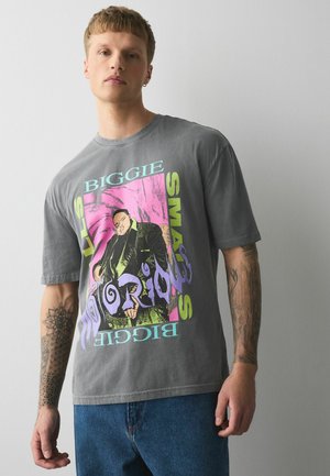 Ung mand med tatoveringer iført en grå Biggie Smalls grafik t-shirt og blå jeans, stående mod en ensartet lysegrå baggrund.