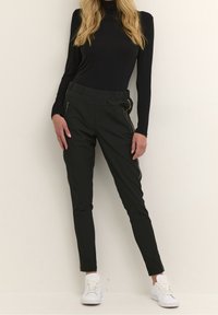 Pantalons ajustés noirs avec une taille élastique. Présente des zips latéraux dorés et des finitions. Modèle portant des baskets blanches, présenté sur un fond clair.