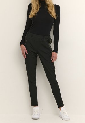 Pantalons ajustés noirs avec une taille élastique. Présente des zips latéraux dorés et des finitions. Modèle portant des baskets blanches, présenté sur un fond clair.