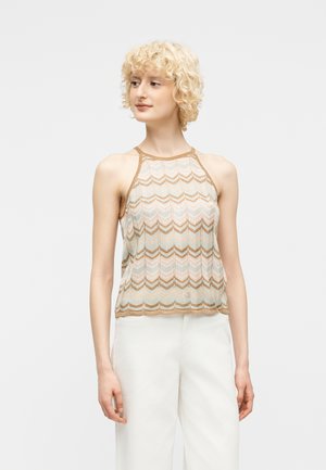 Kvinde med kort krøllet blondt hår iført ærmeløs beige og lyseblå mønstret top og hvide bukser, stående mod en ensfarvet baggrund.