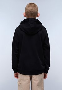 Svart hettegenser med teksturert overflate, ribbestrikkede mansjetter og kant. Barn som har den på, sett bakfra, kombinert med lyse beige bukser.