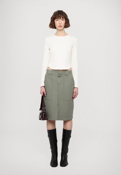 White long-sleeve crop top, green denim skirt with front pockets, black knee-high boots, held burgundy bag with silver accents.