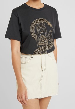 Czarny T-shirt z złotym wężem owiniętym wokół ręki z mistycznymi symbolami, w tym wszechwidzącym okiem, zestawiony z kremową spódnicą jeansową.