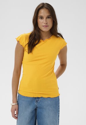 Femme aux longs cheveux bruns portant un haut côtelé jaune à manches courtes et un jean bleu, debout avec une main derrière le dos.