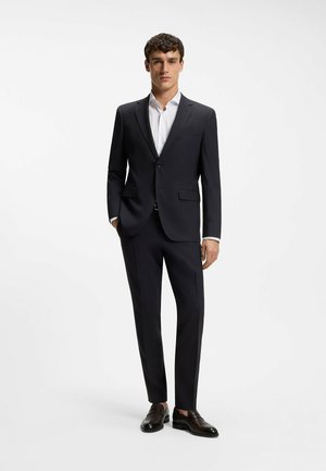 Mann steht und trägt einen schwarzen Anzug mit weißem Hemd und schwarzen Lederschuhen vor einem schlichten weißen Hintergrund.