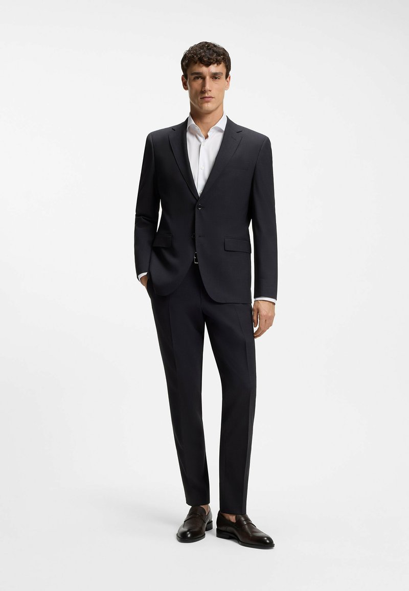 Mężczyzna stojący, ubrany w czarny garnitur, białą koszulę i czarne eleganckie buty na jednolitym białym tle.