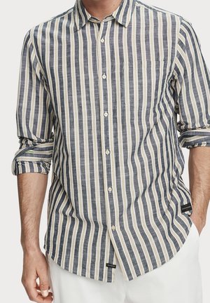 Mężczyzna ubrany w beżową, białą i ciemnoszarą koszulę zapinaną na guziki z pionowymi paskami, z rękawami podwiniętymi do łokci oraz białe spodnie.