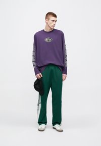 Μωβ πουκάμισο μακρυμάνικο με κεντρικό λογότυπο, σε συνδυασμό με πράσινο αθλητικό παντελόνι με ρίγες στο πλάι και μαύρο καπέλο που κρατιέται στο χέρι.