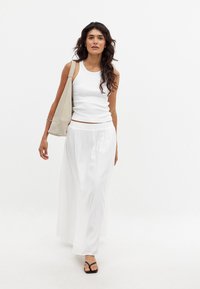 Un débardeur blanc associé à une longue jupe blanche fluide. Un sac à épaule beige, des sandales noires et des cheveux lâchés complètent le look.