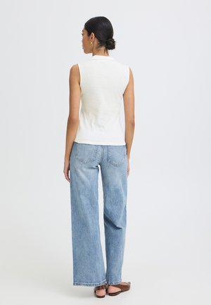 Frau von hinten, trägt ein ärmelloses weißes Oberteil, hellblaue Jeans mit weitem Bein und braune flache Sandalen, steht vor einem schlichten Hintergrund.