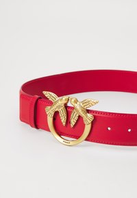 Ceinture en cuir rouge dotée d'une boucle circulaire dorée ornée de deux figures d'oiseaux, détaillées avec des plumes gravées. Ajustable par trous.