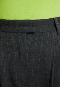 Pantalon gris à fines rayures avec une texture lisse en laine, doté d'un devant plat et d'une ceinture. Le fond est un tissu vert uni.