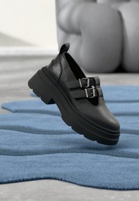 Fekete bőrből készült cipők vastag talppal, dupla csattos díszítéssel és lekerekített orral, egy texturált kék szőnyegen elhelyezve.