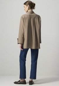 Beige long-sleeve shirt with a collar, relaxed fit, and back pleat. Paired with blue straight-leg jeans and dark loafers.