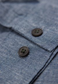 En knappad jeansskjortedel i en texturerad blå tygkvalitet med två svarta knappar som har en korsformad sömnadsdetalj och en kragskant.