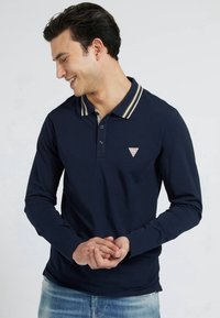 Man smiling and looking down wearing a navy blue long-sleeve polo with striped collar and light blue jeans against a plain background.
