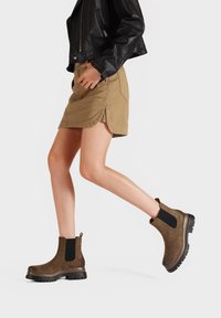 Stivaletti alla caviglia in suede marrone con pannelli laterali in elastico nero e suola in gomma testurizzata, abbinati a una gonna khaki e a una giacca di pelle nera.