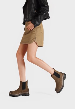Stivaletti alla caviglia in suede marrone con pannelli laterali in elastico nero e suola in gomma testurizzata, abbinati a una gonna khaki e a una giacca di pelle nera.