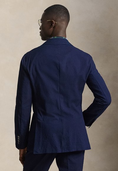 Άνδρας με ναυτικό μπλε υφασμάτινο κοστούμι και γυαλιά, γυρισμένος πλάτη με το ένα χέρι στη μέση, μπροστά σε απλό μπεζ φόντο.