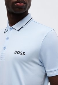 Κοντινό πλάνο ενός ατόμου που φοράει ανοιχτό μπλε πουκαμισάκι πόλο με γιακά με μαύρο περίγραμμα και λογότυπο "BOSS" στο στήθος.