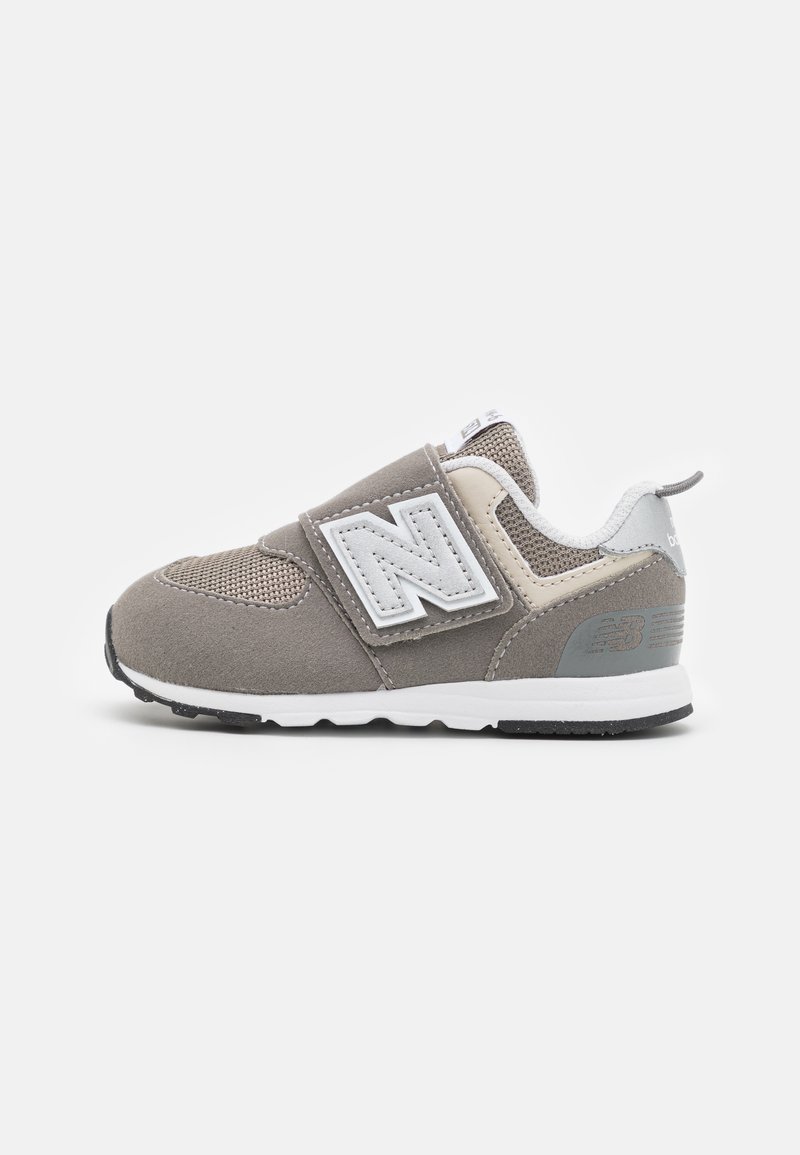 Chaussure de sport grise avec un dessus en maille et en daim, marquage blanc, sangle réglable, semelle intermédiaire blanche et semelle noire crantée.