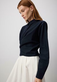 Camicia blu navy cropped con colletto e maniche lunghe, con bottoni frontali e una texture liscia, abbinata a una gonna bianca plissettata.