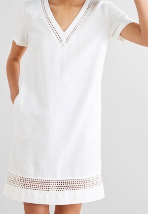 Vestido blanco de manga corta con escote en V y borde con recorte geométrico en el escote y el dobladillo, llevado por una persona con una mano en el bolsillo.