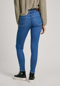 Jeans ajustados de mezclilla de color azul, con bolsillos traseros, cintura de talle medio y un parche de cuero marrón en la cinturilla. Combinados con zapatillas negras.