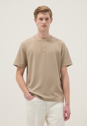 Jonge man, gekleed in een beige korte mouwen henleyshirt met twee knopen en een witte broek, staand met één hand in zijn zak.