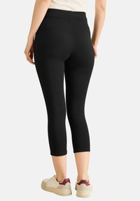 Zwarte capri leggings met een gladde textuur en hoge tailleband, gecombineerd met beige sneakers. Minimale vormgeving zonder zichtbare patronen of accenten.