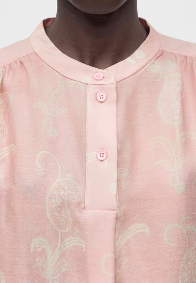 Άτομο με σκούρο δέρμα που φοράει ανοιχτό ροζ μπλούζα με μανδαρινέ γιακά, τρία ροζ κουμπιά και διακριτικά λουλουδάτα σχέδια.