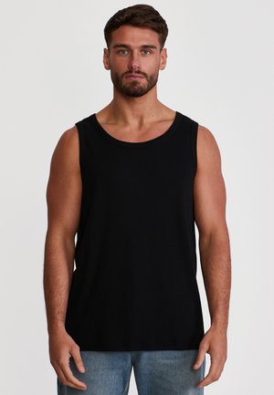 Mężczyzna z brodą w czarnej koszulce bez rękawów i niebieskich dżinsach, stojący na tle jasnego, jednolitego tła.