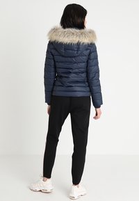 Námořnicky modrá péřová bunda s kapucí lemovanou kožešinou, prošívaným vzorem a žebrovanými manžetami. Nošena s černými kalhotami a bílými sportovními botami.