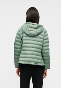 Άτομο με ανοιχτοπράσινο μπουφάν με κουκούλα και μαύρο παντελόνι, που φαίνεται από πίσω πάνω σε απλό φόντο.