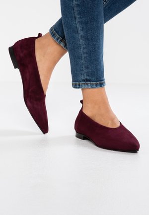 Kvinde iført bordeaux ruskind spidse flats kombineret med optrukne blå skinny jeans på hvidt gulv.