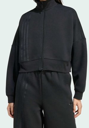 Personne portant une veste noire zippée et un pantalon assorti avec un détail discret à trois bandes sur les côtés gauches.