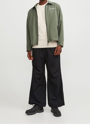 Man met een olijfgroene jas met rits over een beige shirt, zwarte wijd uitlopende broek en zwarte wandelschoenen, staand tegen een effen lichte achtergrond.